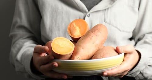 Woman with bowl of raw sweet potatoes on light background, closeup