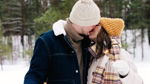Happy young couple kissing with sparklers in snowy winter forest on christmas holidays
