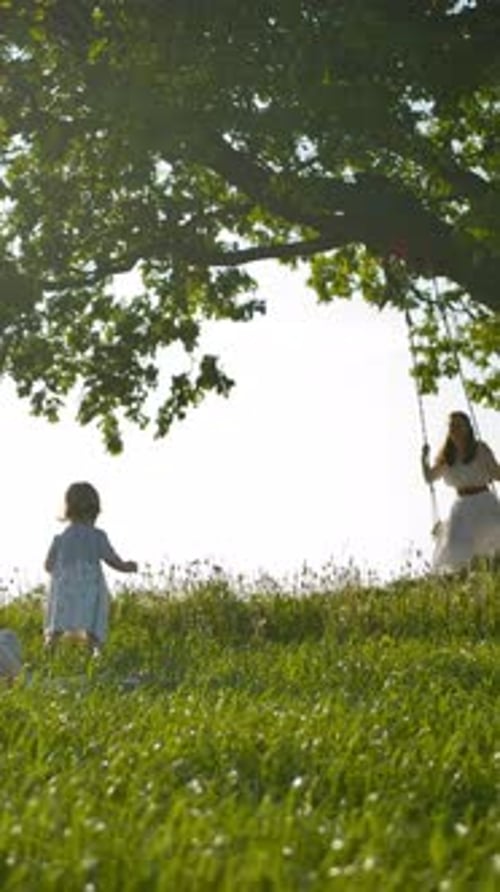 Vertical of Two Little Sister Girls Run Across a Clean Green Field to Their Mother Swinging on a