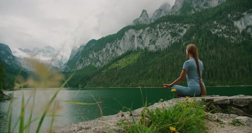 Woman meditates on a rocky ledge overlooking a tranquil lake and towering peaks