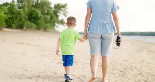 Mother Holding Hand with Little Son Walking Together on Sandy Beach at Summer