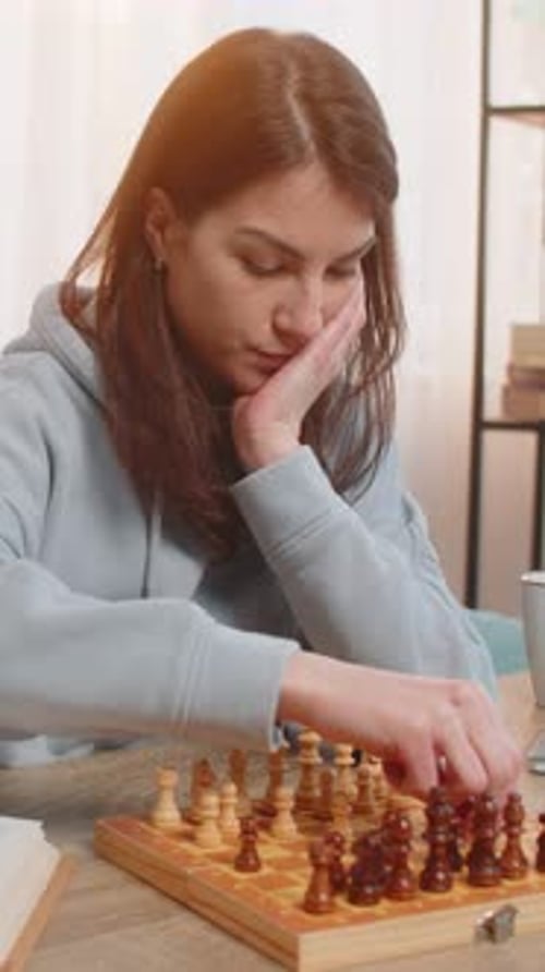 Young Woman Playing Chess Alone Arranging Pieces at Table on Sofa with Book Lying Nearby at Home