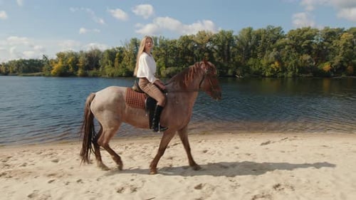Caucasian Sportswoman Galloping at Sandy Lake Beach with Green Forest Around