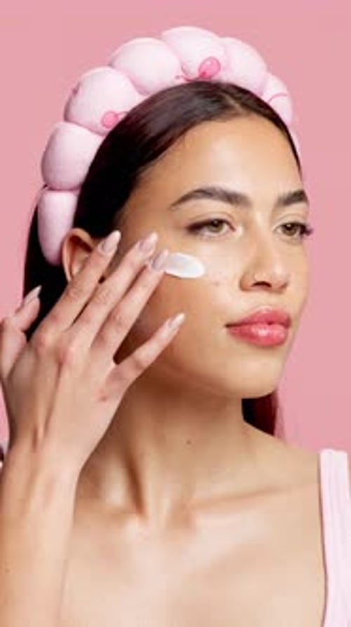 Woman, cream and skincare with headband in studio, thinking and application on pink background