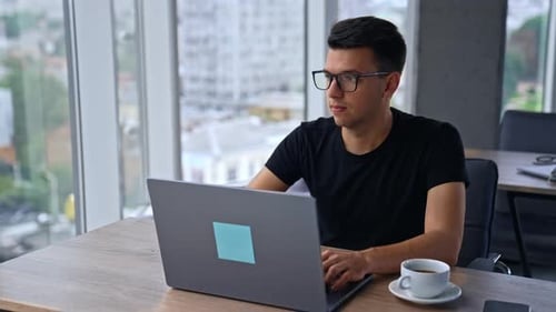 Pensive young man in glasses and black t-shirt sits in the office in front of laptop.