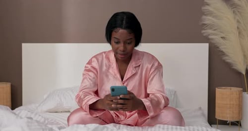 Mulher feliz e sonolenta sorridente senta-se de pijama na cama do quarto e segura um telefone nas mãos que ela envia