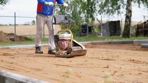 Compacteur à plaque d'opération Worker sur un terrain de construction sablonneux à côté d'un site bordé de trottoir pendant