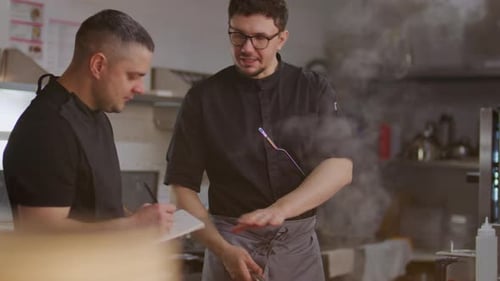 Chef Explaining Recipe to Apprentice Taking Notes in Restaurant Kitchen