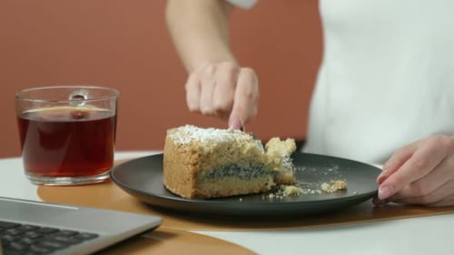 Woman Eats Cake with Tea Next to Laptop