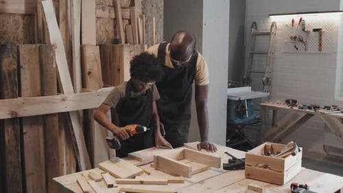 Child Making Holes in Plank with Drill while Father Helping Him in Workshop