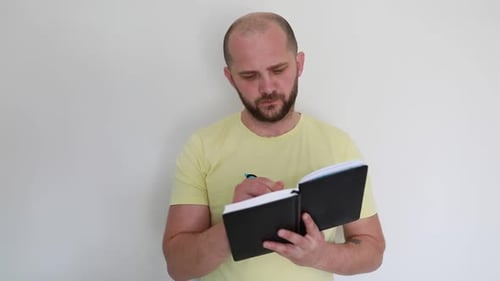 Man in Yellow Shirt Intently Reading a Black Notebook Indoors