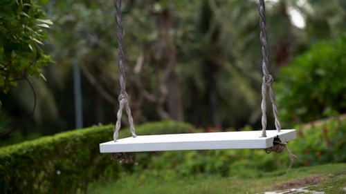 A White Swing Swings Against the Backdrop of a Green Garden