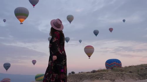Young Female Tourist Watching Hot Air Balloons Flying Over Goreme At Sunrise In Cappadocia, Turkey.