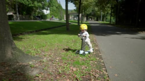 Toddler riding scooter on park path wearing yellow helmet, slows down and stops after rolling onto