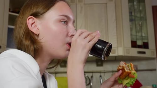 Teenage Girl Eating Burger and Drinking Drink at Home