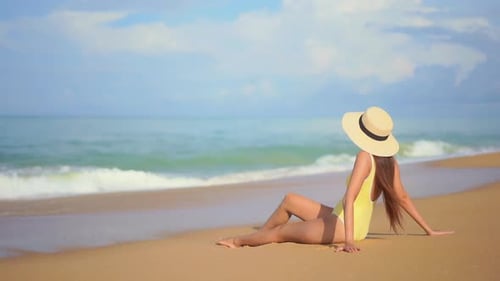 Einsame Frau in Badeanzug und Sommerhut sitzt auf Strandsand und blickt auf S auf die Wellen des tropischen Meeres