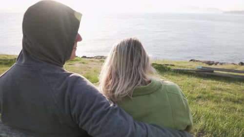 Adult couple sitting on bench with ocean view during sunset, back view