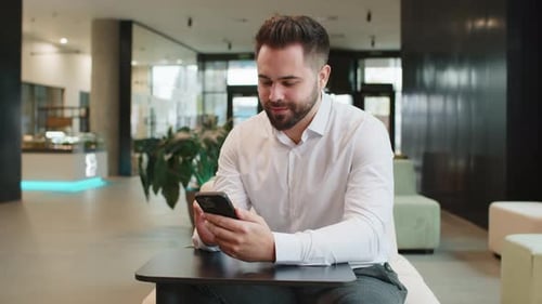 Smiling Caucasian Businessman Using Smartphone Typing Text Messages Browsing News in Office Lobby