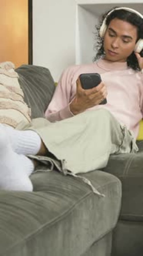 Vertical video: Reclining nonbinary adult scrolling phone at home, adjusting headset to relax
