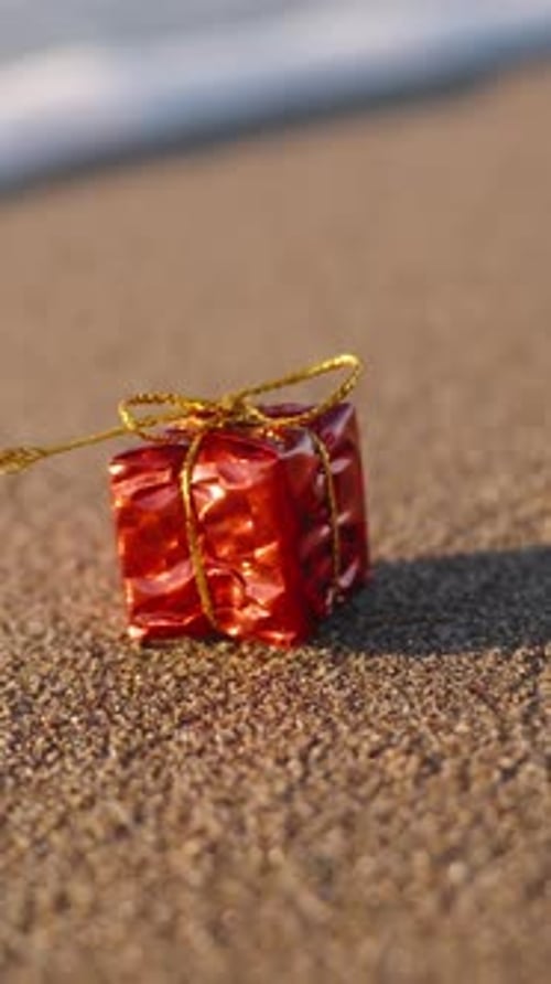 Vertical video. micro gift box in a red wrapper with a golden bow, on a sandy beach by the sea