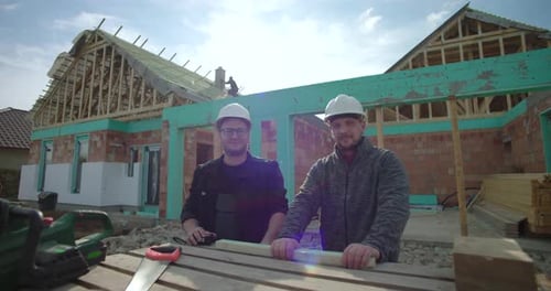 Dos trabajadores de la construcción posando con herramientas frente a una casa de madera sin terminar, trabajo en equipo