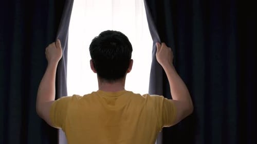 Man stands at window and opens curtains, back view.