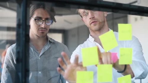 Junge Geschäftsleute in formeller Kleidung planen die Arbeit im Büro mit Papieraufklebern auf dem Glas