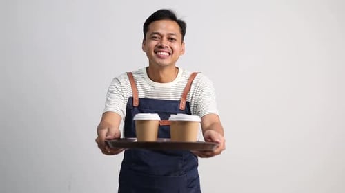 A male coffee shop waiter wearing an apron while carrying a tray containing paper cups of coffee