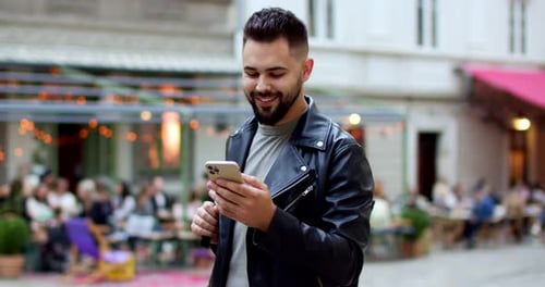 Cheerful Young Handsome Man in Black Leather Jacket Standing at Street and Texting Message on
