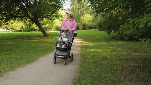 Young Mother Walking in the Summer Park with Her Young Daughter in a Baby Carriage Outdoors