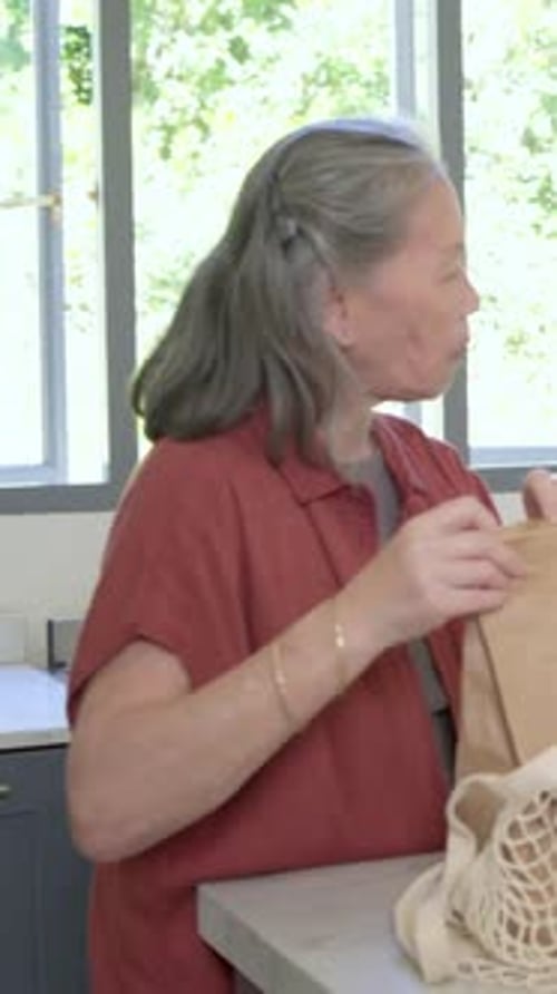 Vídeo vertical: Llegando a una mujer mayor con una blusa oxidada desempacando una bolsa de malla y bolsas marrones en el mostrador