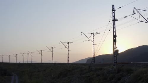 Silhouette einer leeren Eisenbahn mit Hochspannungsleitungen und Sonne, die sich hinter einem Hügel versteckt, bei Sonnenuntergang