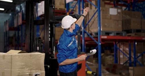 Warehouse supervisor in blue uniform holding tablet and pointing at upper rack while inspecting