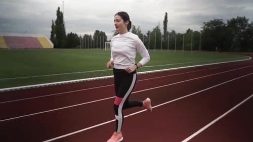 Young woman is running in the stadium. woman with white sweater and black tights. Camera follows run