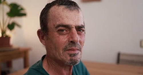 Senior man with gray hair and beard looks seriously into the camera at home
