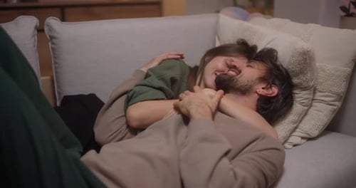 Happy Brunette Girl Hugging with Her Boyfriend While Lying on the Sofa in the Evening at Home