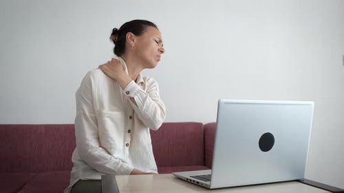 Woman Massaging Sore Shoulder at Laptop Indoors