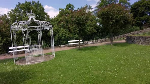 A white wedding gazebo in the middle of a green garden. Castle garden with paths and benches in sunn