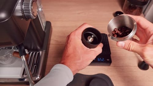 Top Down View Of Barista Pouring Fresh Ground Coffee From Metal Cup Into Manual Grinder Hopper On