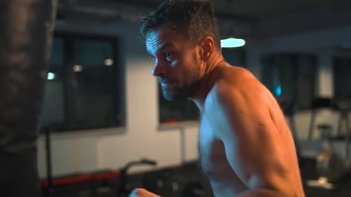 Alone Boxer is Working Out Blows on a Punching Bag in a Gym in Evening