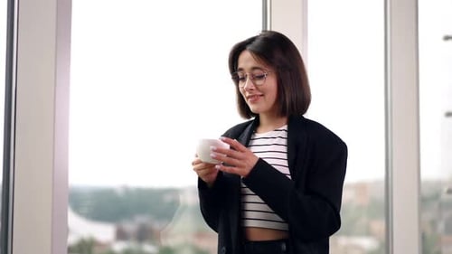 Caucasian lady standing at the window. Woman drinks coffee looking at the window. Low angle view.