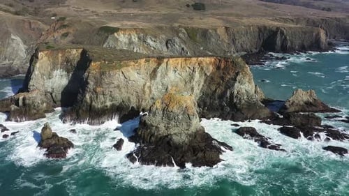 Aerial view of ocean waves crashing against large cliff rocks with slow pan up and out.