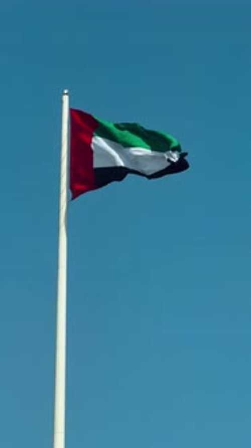 A close-up shot of the UAE national flag waving boldly atop a tall flagpole, set against a clear