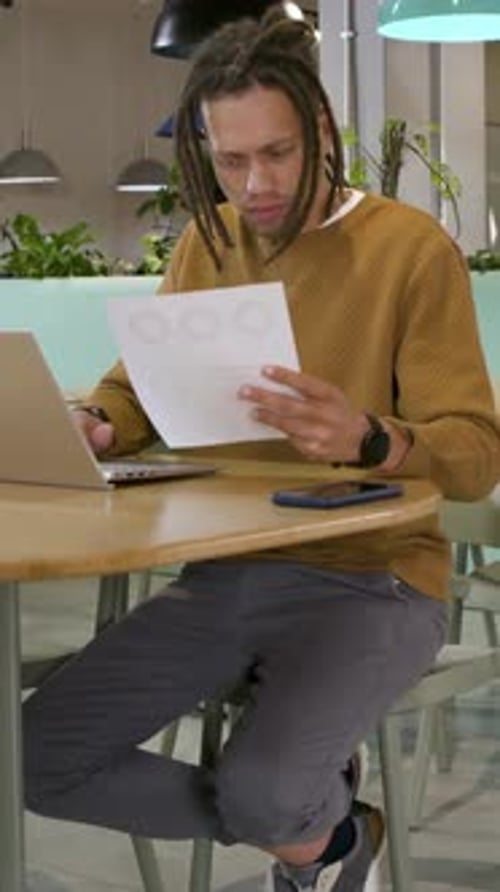 Vertical video: Typing man with dreads working through charts on laptop at work table tapping phone