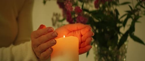Les mains se réchauffent autour d'une bougie avec des fleurs en arrière-plan à l'intérieur