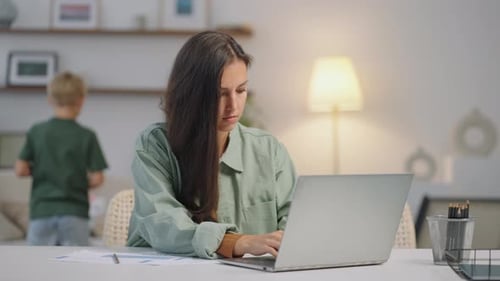 A Business Woman Works Remotely at Home on a Laptop While a Family Plays in the Background