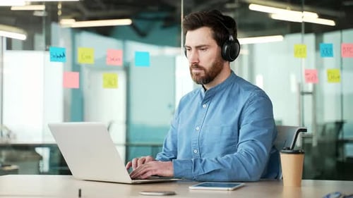A focused IT specialist in wireless headphones works on a laptop sitting at a workplace