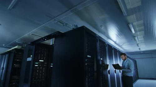 Bearded IT Specialist in Glasses is Working on Laptop in Data Center while Standing Before Server R