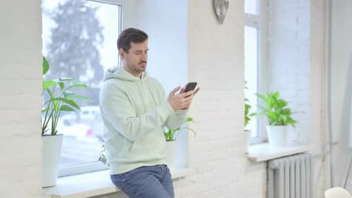 Young Man using Smartphone while Standing Near Window