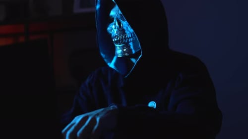 Hacker con máscara facial y sudadera con capucha que usa una computadora portátil en la oscuridad ilustrando el robo de identidad y lo digital